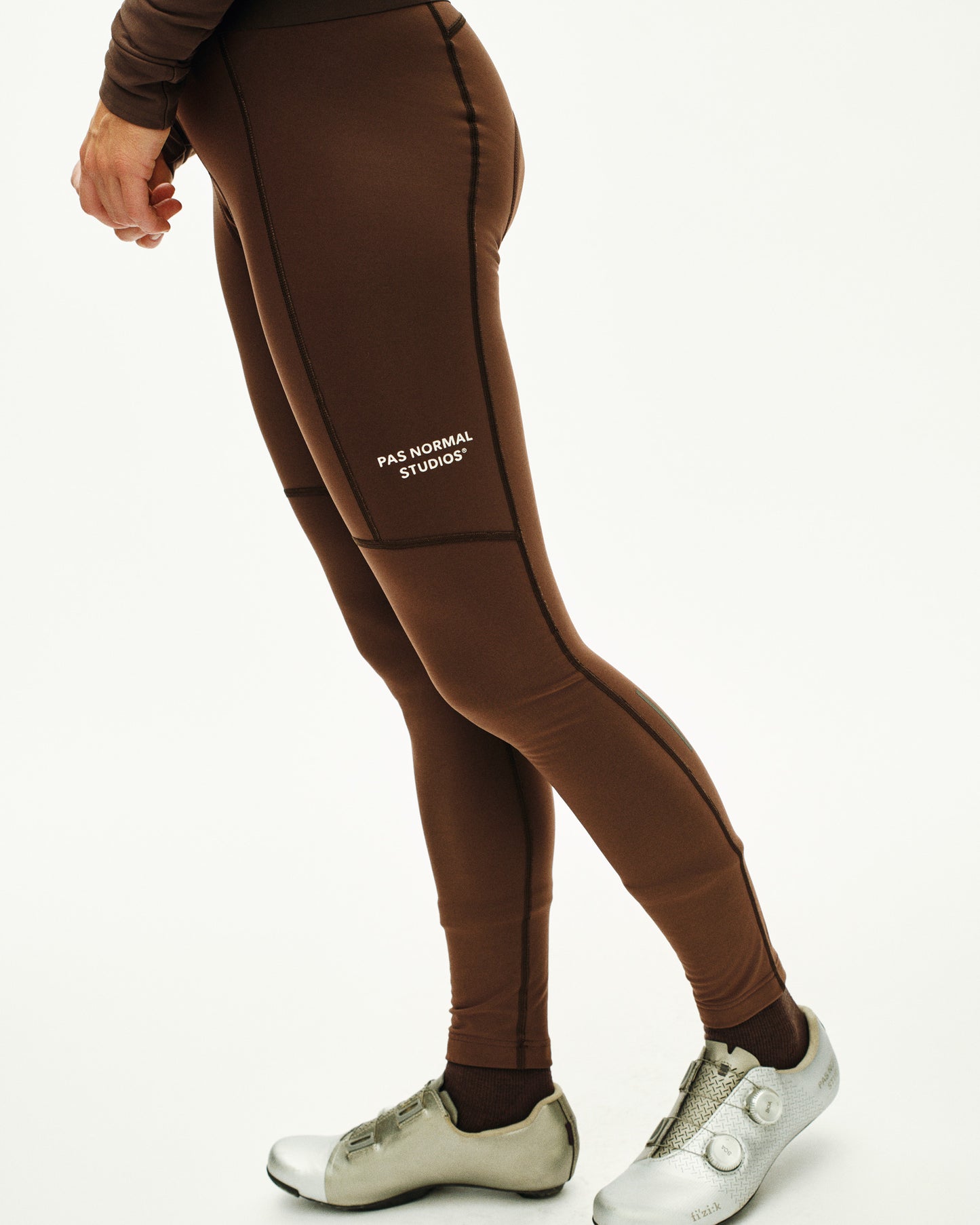Brown leggings with 'Pas Normal Studios' logo worn by a person on a white background