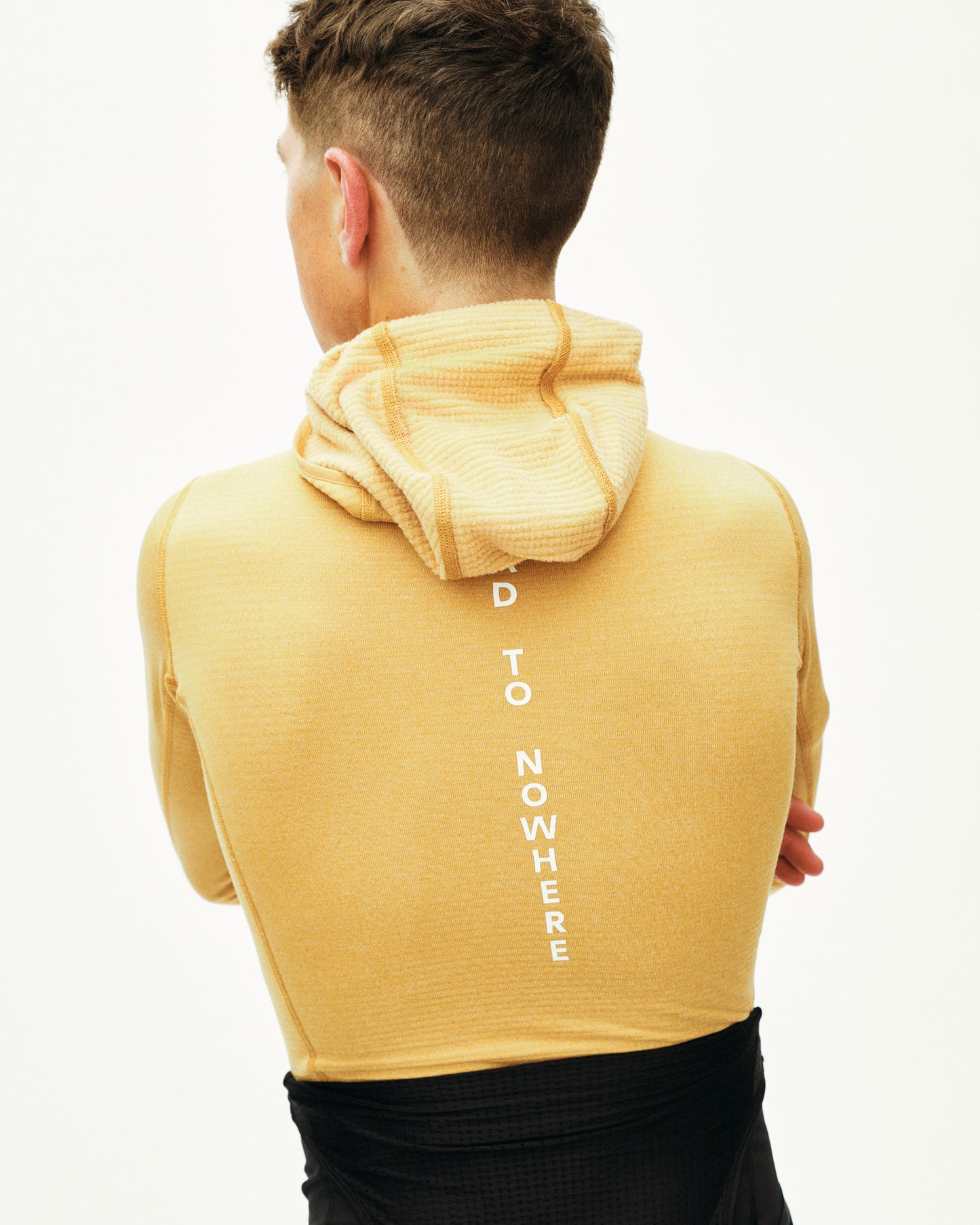 Person wearing a mustard yellow hoodie with text on the back, standing against a white background