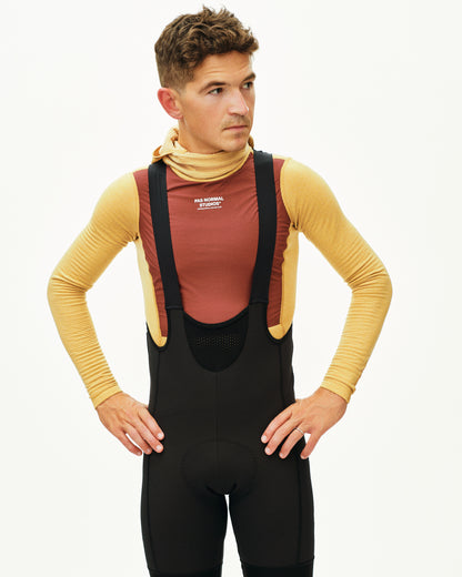 Man wearing a brown shirt, black bib overalls, and a beige turtleneck against a white background