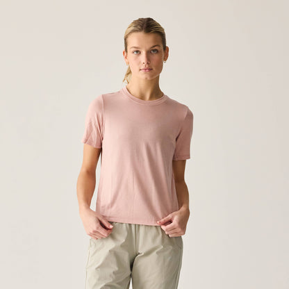 A woman wearing a short-sleeved pink t-shirt and beige pants standing against a plain background.