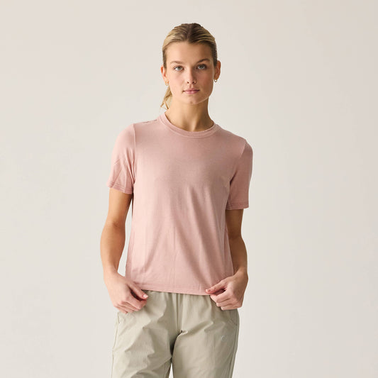A woman wearing a short-sleeved pink t-shirt and beige pants standing against a plain background.